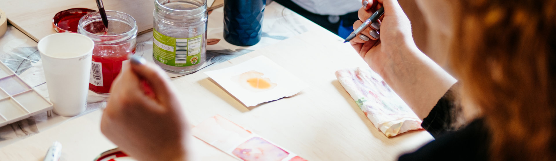 Eine Person sitzt an einem Tisch und malt mit Aquarellfarben auf einem Blatt Papier.