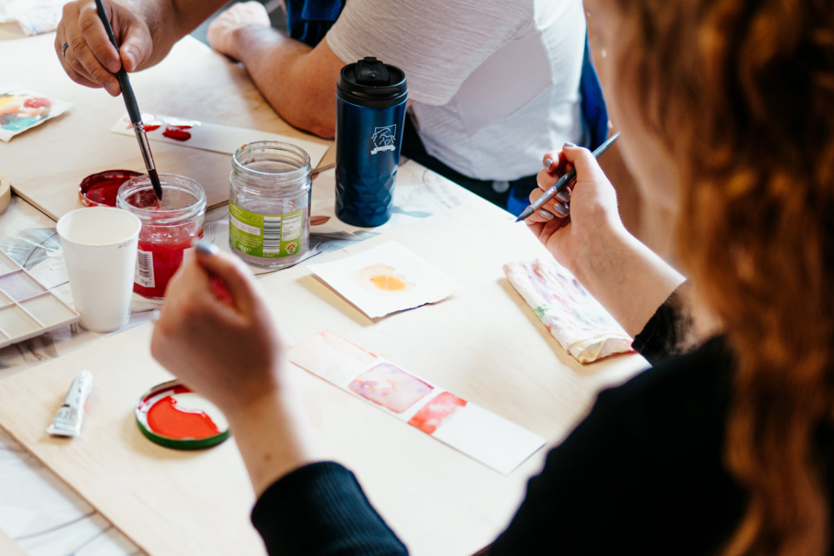 Eine Person sitzt an einem Tisch und malt mit Aquarellfarben auf einem Blatt Papier.